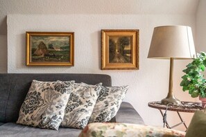 A room with a grey sofa, two patterned cushions, a lamp on a side table, and two framed paintings on the wall. - Holiday apartment with 1 bedroom (Born am Darß)