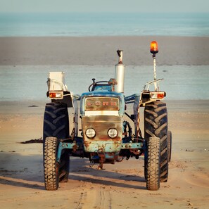 Ein alter Traktor steht auf einem sandigen Strand, im Hintergrund das Meer.