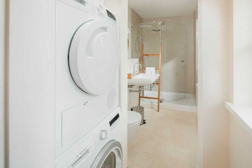 Un espace buanderie équipé d’un lave-linge et d’un sèche-linge, une douche avec une porte en verre et un lavabo avec un tabouret en bois.