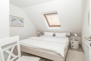 Ein Schlafzimmer mit geneigter Decke, einem Oberlicht, einem Bett mit weißer Bettwäsche, einem Nachttisch und einem gerahmten Bild an der Wand.
