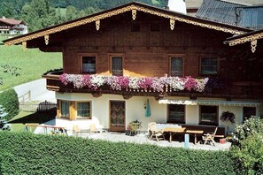 A two-story wooden chalet with a balcony adorned with flowers, a dining area with outdoor seating, and a view of a green hillside. - Holiday apartment with 2 bedrooms (Schwendau\/Mayrhofen)