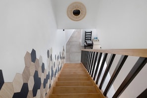 Una escalera con un muro de diseño geométrico, barandilla de madera y una silla con un libro sobre una mesa en la parte superior.