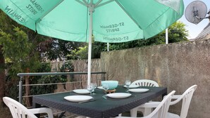 Un espacio de comedor al aire libre con una mesa preparada para cuatro personas, rodeada de sillas blancas, bajo un paraguas verde.