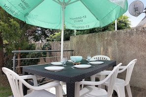 Un espacio de comedor al aire libre con una mesa preparada para cuatro personas, rodeada de sillas blancas, bajo un paraguas verde.