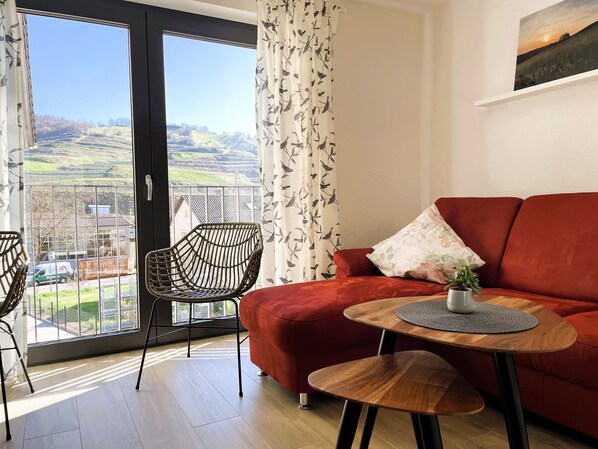 Uma sala de estar moderna com um sofá vermelho, uma mesa de centro de madeira e uma cadeira de vime. Uma janela grande oferece vista para uma paisagem verde.