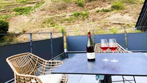 Een terras met een tafel, een rieten stoel en een fles wijn met glazen.