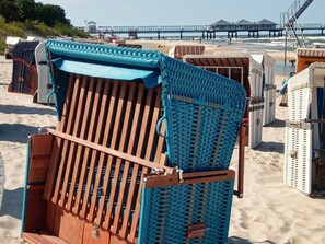 Strandstühle mit einem gewobenen Rücken und einer Holzsitzecke, auf einem Sandstrand mit einem Pier im Hintergrund.
