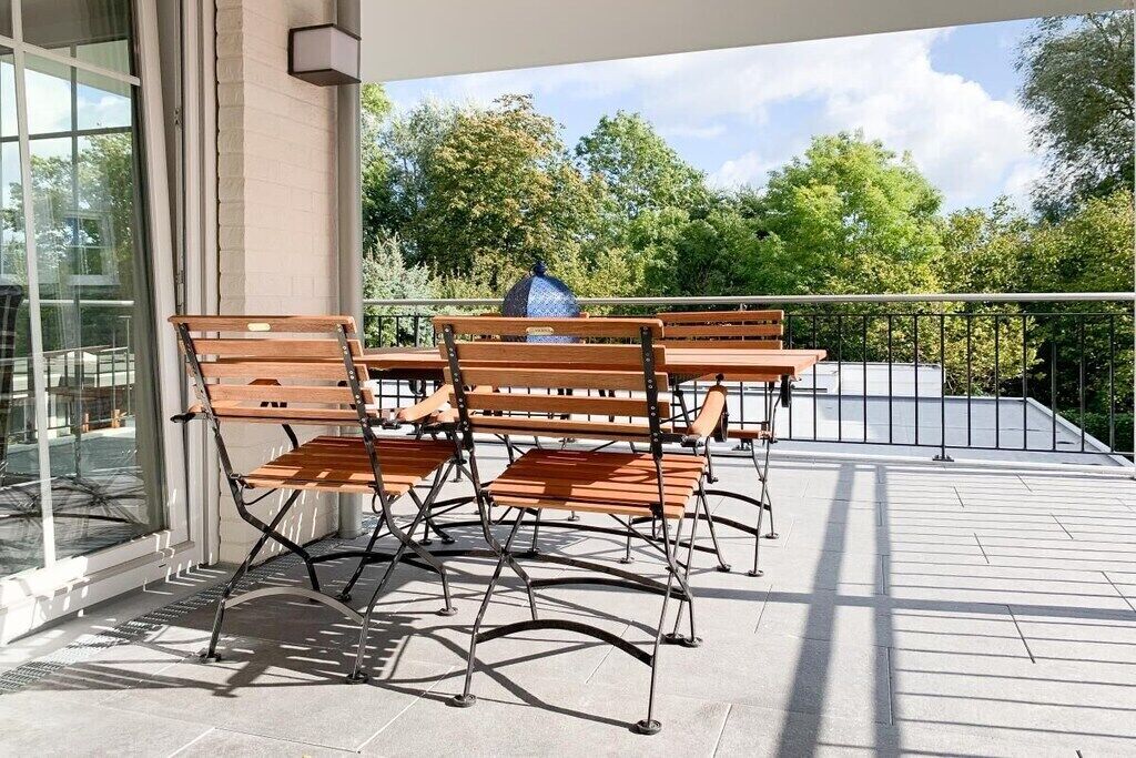 A balcony with outdoor furniture, including wooden benches and a table, and a view of trees and sky.