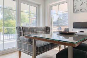 A modern living room with a glass-top table, plaid sofa, and a large window with a view of a residential area. - Holiday apartment with 2 bedrooms (Timmendorfer Strand)