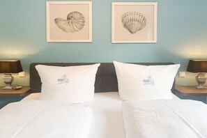 Un lit double avec une literie blanche et des oreillers assortis, deux œuvres d’art représentant des coquillages encadrées au mur, et des lampes de chevet de chaque côté.