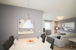 A modern living room with a dining area, a white table with a bowl of fruit, a white sofa, and a hanging lamp. - Holiday apartment with 1 bedroom (Scharbeutz)