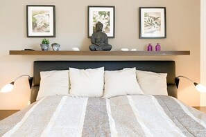 Een bed met hoofdbord, twee bedlampen en een plank erboven met een beeldje en ingelijste foto's.