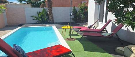 Piscina con sillones de descanso y una mesa, rodeada de vegetación y un muro blanco.