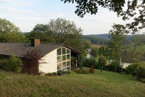 A house with a sloped roof, a large window, and a garden with trees and grass. - Holiday home in Bürberg with swimming pool (Bürberg)