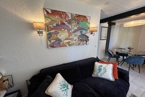 A living room with a black sofa, two cushions, a wall-mounted fish painting, and a dining area with a glass table and chairs. - Holiday apartment in Lumio with swimming pool (Lumio)