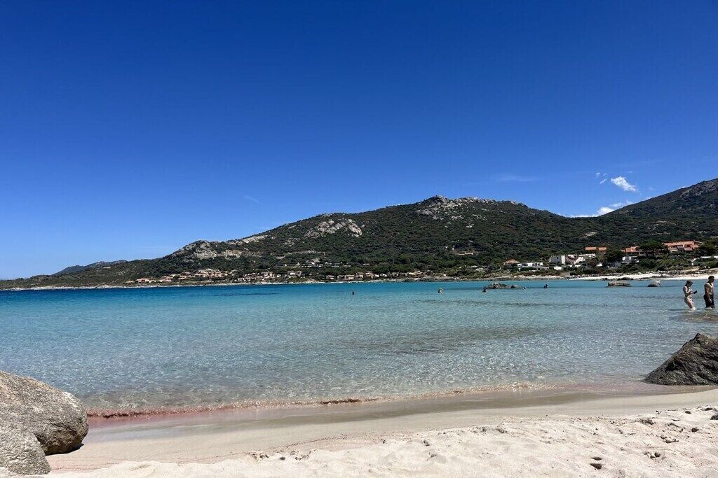 En strand med klart vatten, en sandstrand och ett berg i bakgrunden.