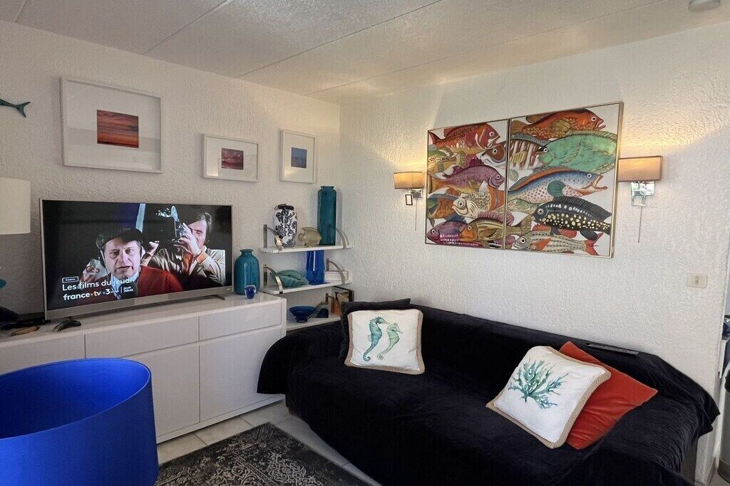 Un soggiorno con TV a schermo piatto, un divano nero, cuscini decorativi, un quadro di pesci appeso al muro e una piccola mensola con oggetti.