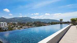 A clear blue infinity pool with a view of a mountainous landscape and buildings in the background. - 2BR family suite near Boat Lagoon (BangTao)