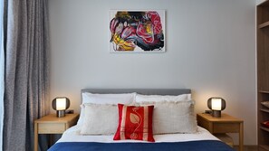 A bedroom with a bed, two bedside tables with lamps, a grey curtain, and a colorful abstract painting on the wall. - 2BR family suite near Boat Lagoon (BangTao)