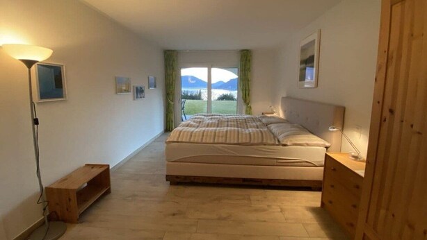 A bedroom with a bed, a bedside table, a lamp, a wooden bench, and a view of the outdoors. - Casa dell' Orso || Morcote (Morcote)
