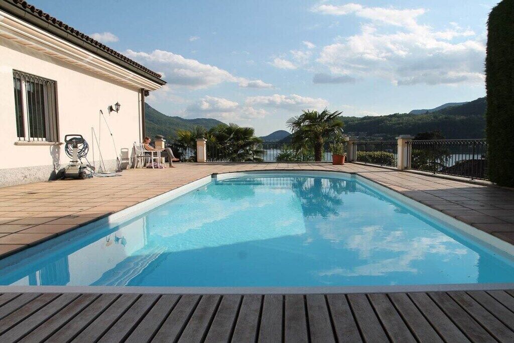 A swimming pool with a wooden deck, a white building with a tiled roof, and a mountainous landscape in the background.