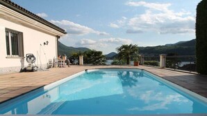 A swimming pool with a wooden deck, a white building with a tiled roof, and a mountainous landscape in the background. - Casa dell' Orso || Morcote (Morcote)