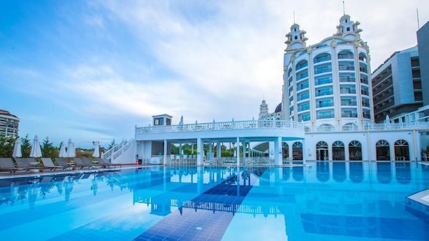 A large swimming pool with a white building featuring arched windows and a balcony in the background. - J'adore Deluxe Hotel & Spa All Inclusive (Manavgat)
