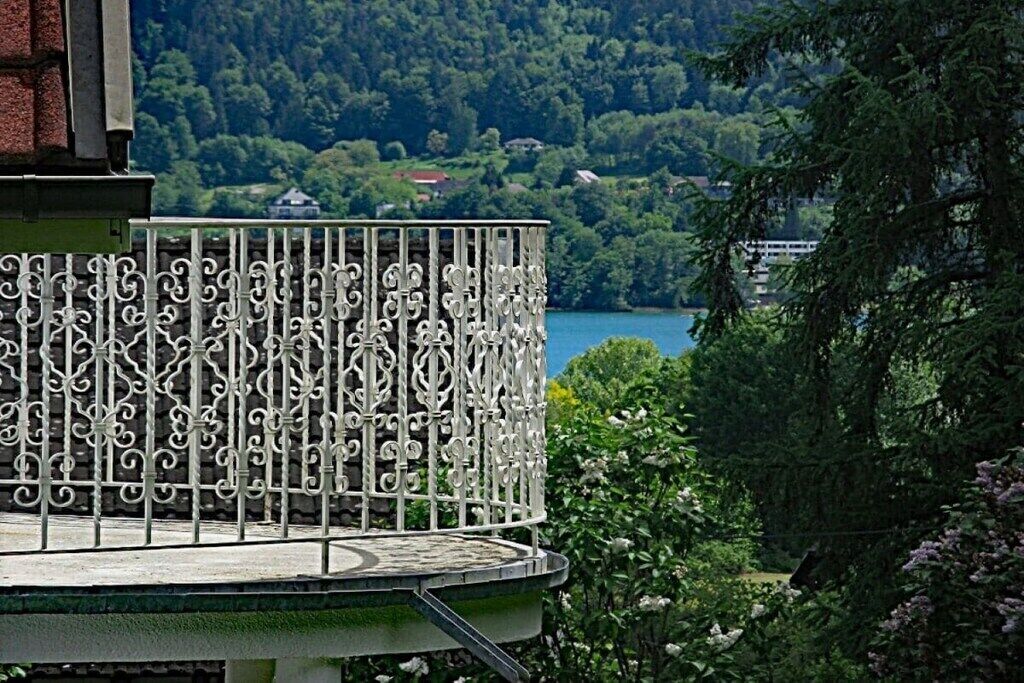 Un balcón con barandillas de metal decoradas, con vista a un lago y una zona boscosa.