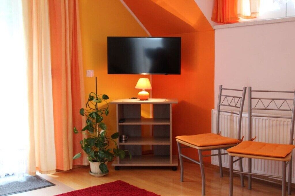 A room with a television mounted on the wall, a small table with a lamp, two chairs, a radiator, and a potted plant.
