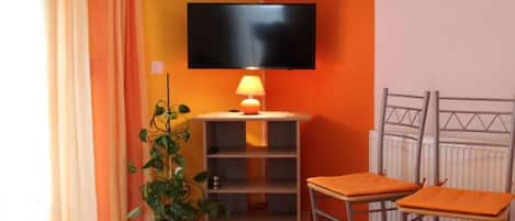 A room with a television mounted on the wall, a small table with a lamp, two chairs, a radiator, and a potted plant.
