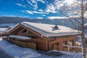 A wooden cabin with a sloped roof, surrounded by snow-covered ground and hills.