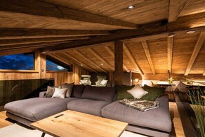 A cozy loft-style room with a wooden ceiling, a sectional sofa, a wooden coffee table, and a window with a view.