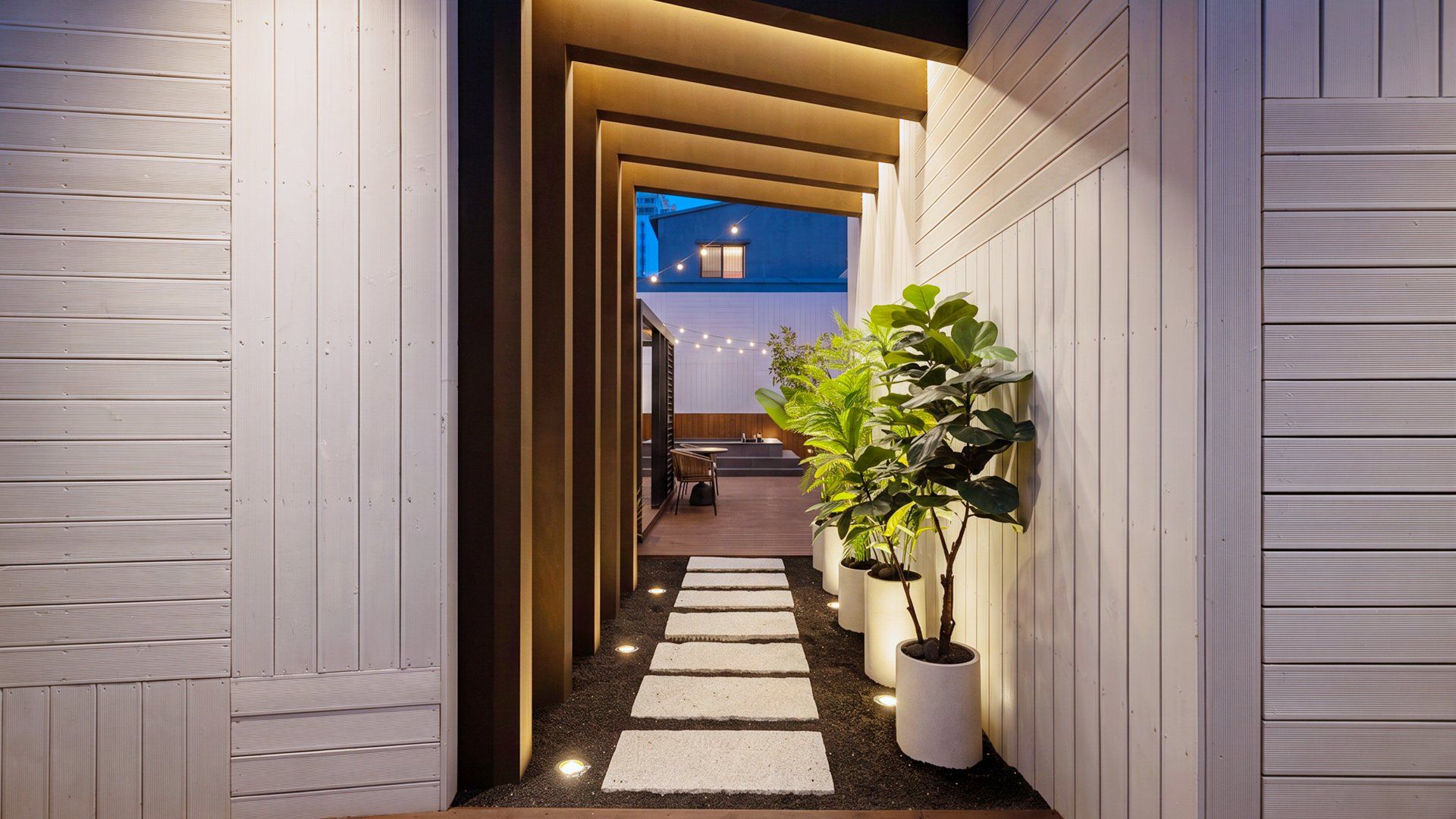 Un couloir étroit doté d’un plancher en bois, de plantes en pot et offrant une vue sur une pièce dont un mur est bleu et qui possède une fenêtre.