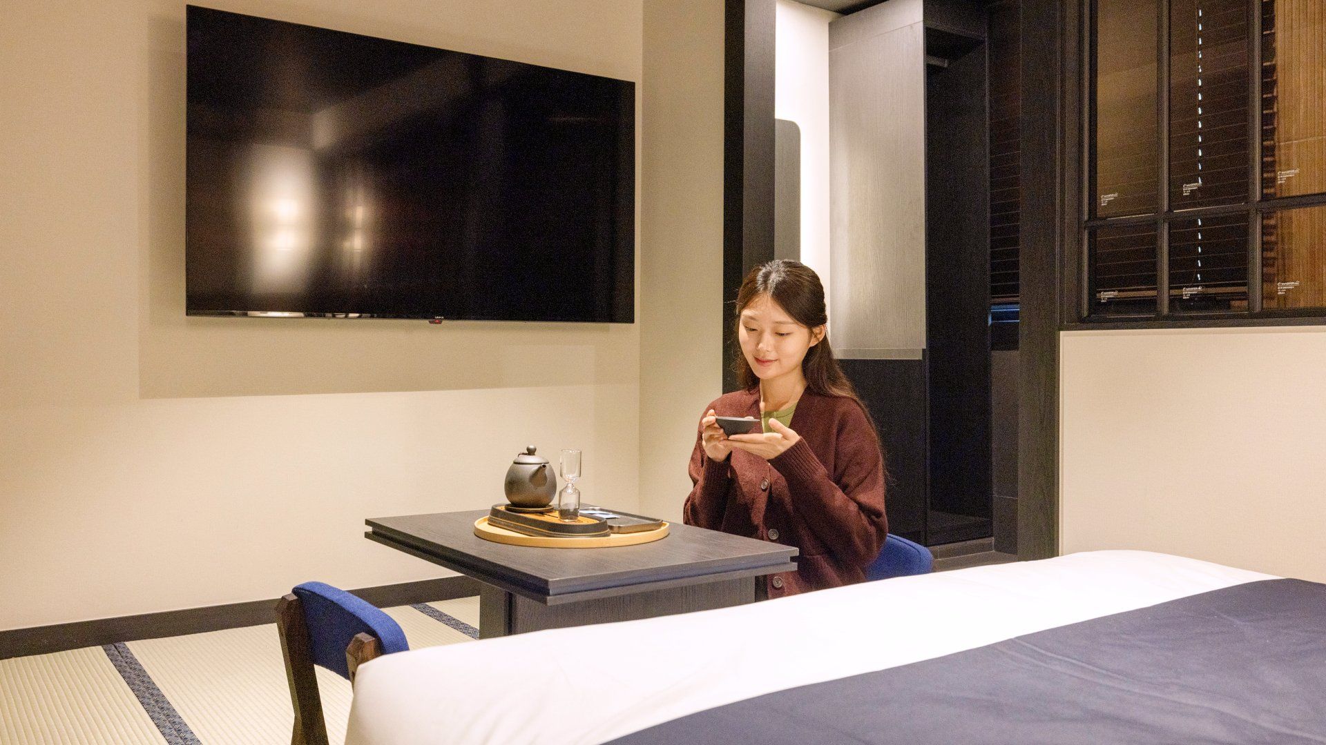 A hotel room with a bed, a table with a teapot and cup, a chair, and a flat-screen TV mounted on the wall.