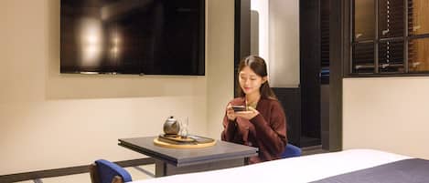 A hotel room with a bed, a table with a teapot and cup, a chair, and a flat-screen TV mounted on the wall.