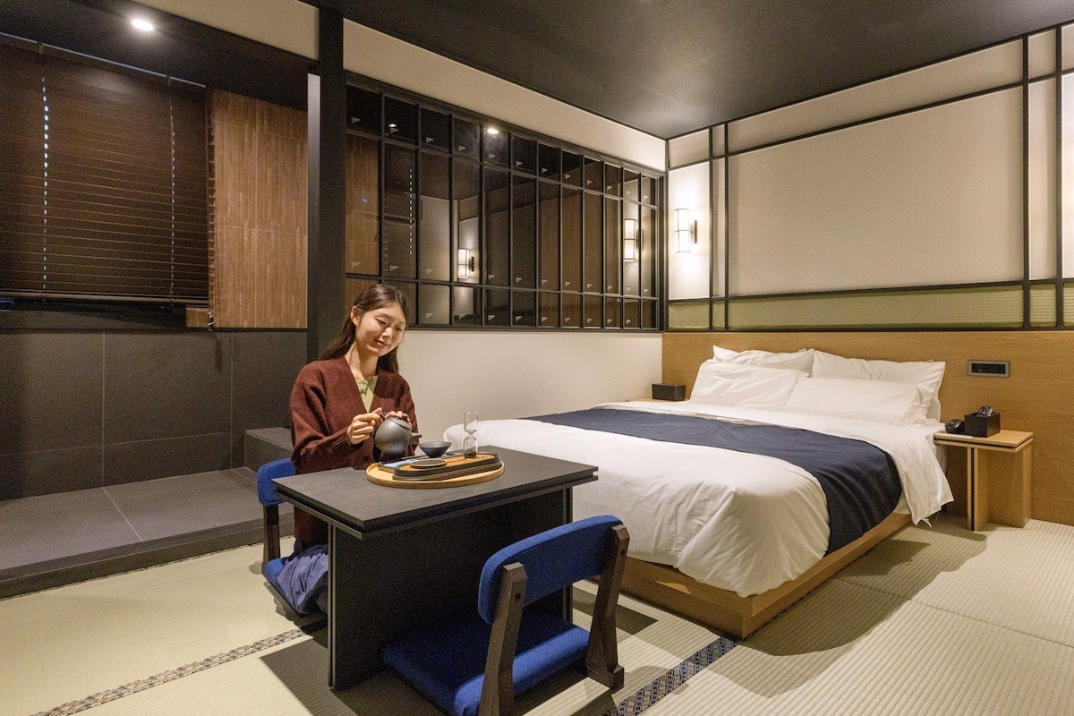 Una habitación de hotel con una cama, una mesita con tetera, una silla y una mesita de noche.
