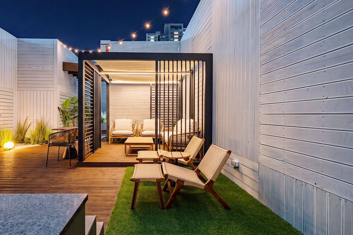 Un patio exterior con terraza de madera, zona de estar y vista del horizonte de la ciudad al caer la noche.