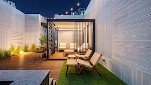 Un patio exterior con terraza de madera, zona de estar y vista del horizonte de la ciudad al caer la noche.