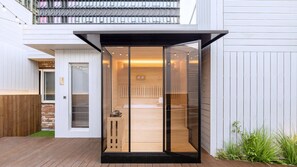 Una sauna moderna, con paredes de vidrio y un interior de madera, junto a un edificio blanco con terraza de madera.