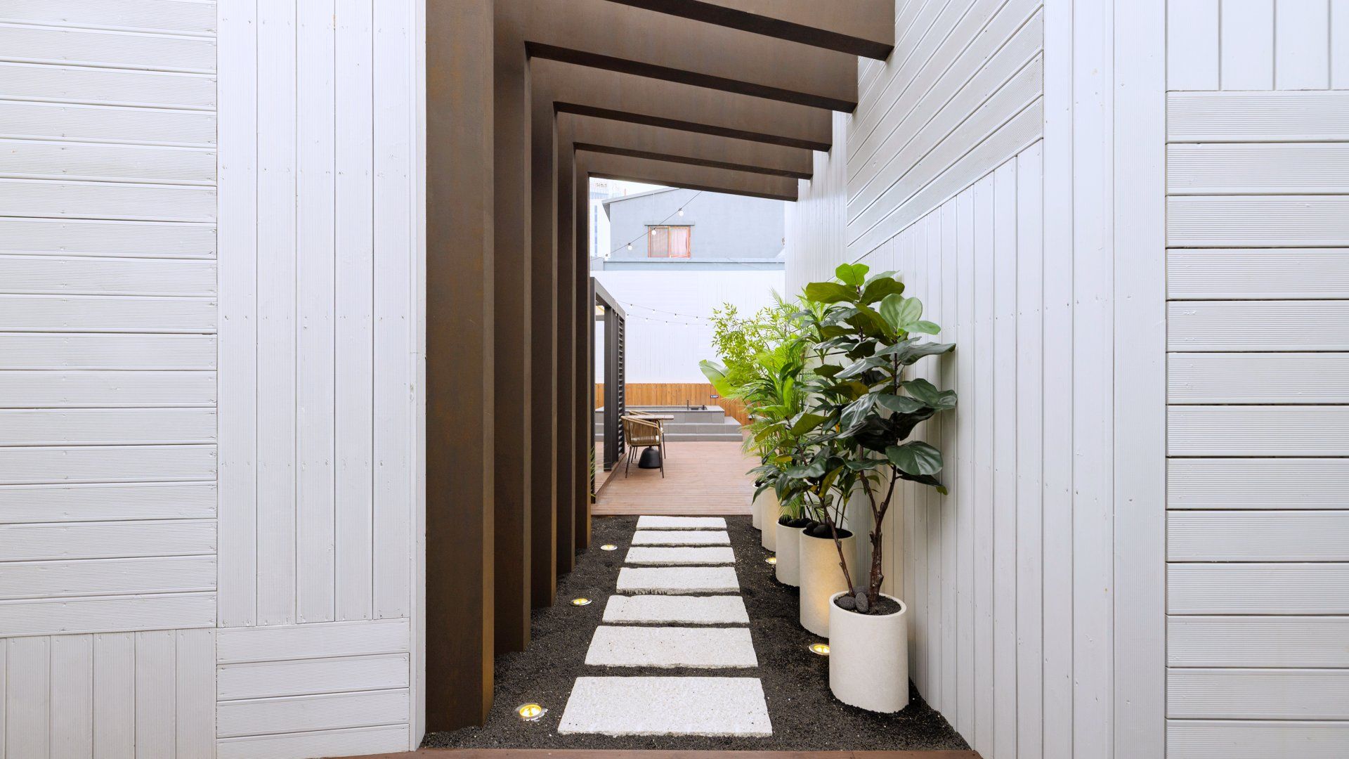 Un étroit sentier, ponctué de pierres plates servant de marchepieds, mène à une terrasse en bois, bordée de murs blancs et de plantes en pot.