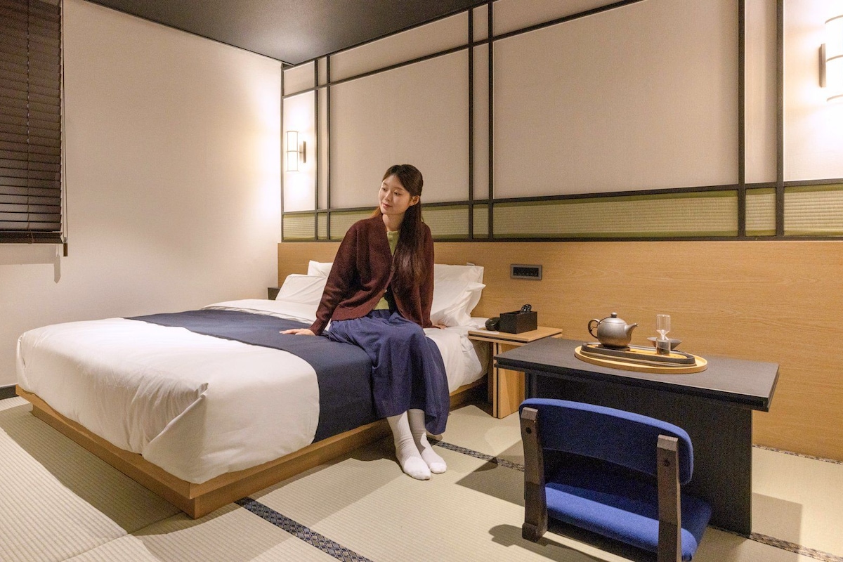 Una habitación de hotel con una cama, un escritorio con tetera y taza, y una silla.