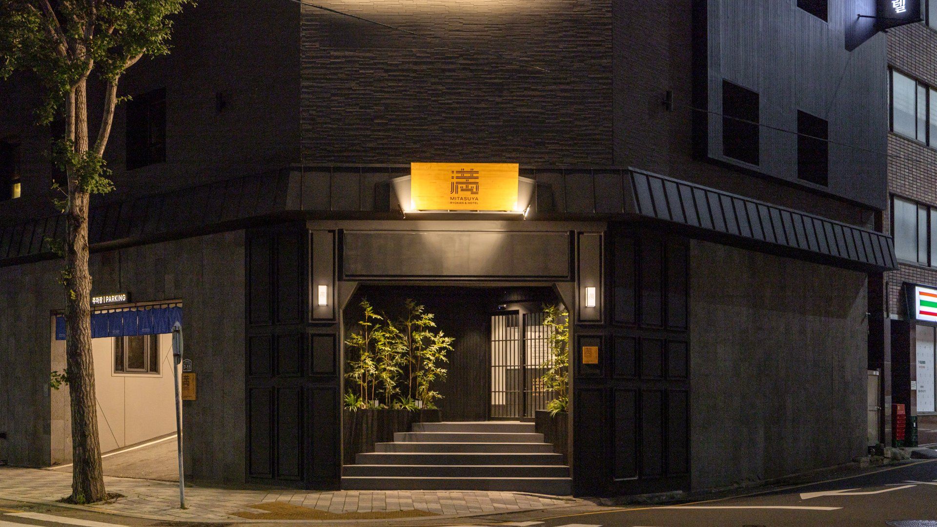 A building with a sign for 'MITSUYA RYOKAN & HOTEL' at the entrance, a staircase leading up, and a parking sign to the left.