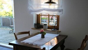 A dining area with a wooden table, chairs, a vase with a flower, a window with a view, and a small shelf with a basket and a device. - Holiday apartment with 1 bedroom (Milda, Deutschland)