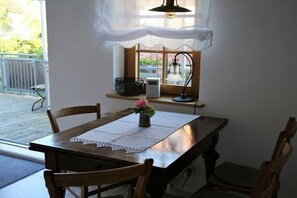 Un coin repas avec une table en bois, des chaises, un vase avec une fleur, une fenêtre donnant sur un paysage et une petite étagère avec un panier et un appareil.