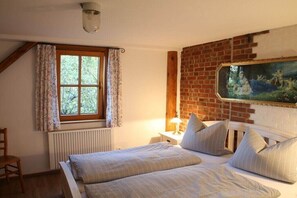 A bedroom with a bed, a window with curtains, a radiator, a nightstand with a lamp, and a painting on the wall. - Holiday apartment with 1 bedroom (Milda, Deutschland)