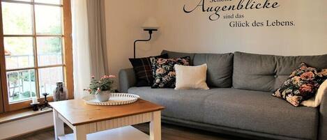 A living room with a grey sofa, a wooden coffee table, and a wall with a German quote.