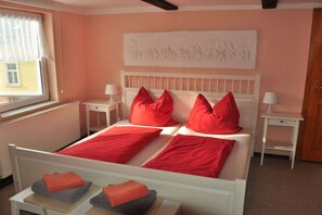 Une chambre avec un lit blanc, des oreillers rouges et une tête de lit blanche. On y trouve une fenêtre avec des rideaux blancs, un radiateur et une œuvre d’art accrochée au mur.