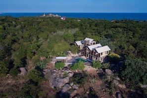 A large house with a pool surrounded by rocks and trees, overlooking a coastline.