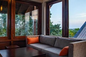 A room with a sofa, a table, and large windows offering a view of greenery.