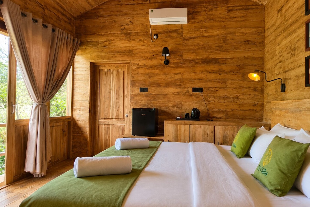 A wooden cabin room with a bed, a small kitchenette, a door, and a window with curtains.